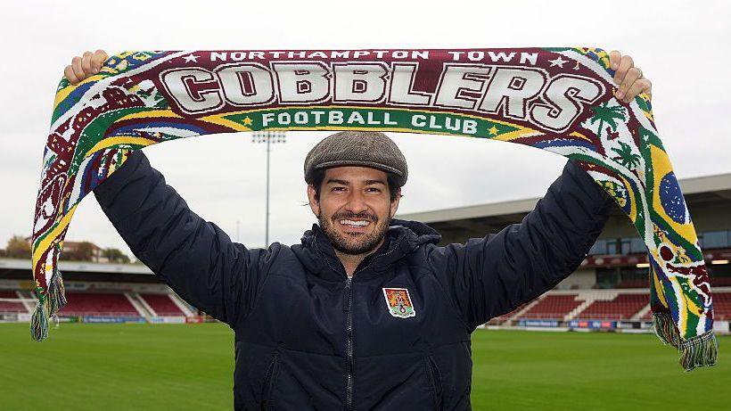 Alexandre Pato holding up a maroon, yellow and blue scarf with the words Northampton Town Football Club and Cobblers on it, smiling towards the camera while wearing a grey flat cap and a coat with the Northampton Town crest on it on the pitch at Sixfields.