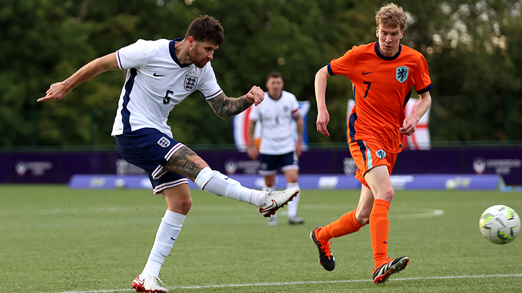 Harry Baker in a white England shirt and blue shorts strikes a ball as a Dutch defender - in an all orange kit - watches on.