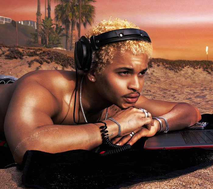 Young man with blonde curly hair wearing headphones, lying on the beach at sunset, focused on a laptop nearby.