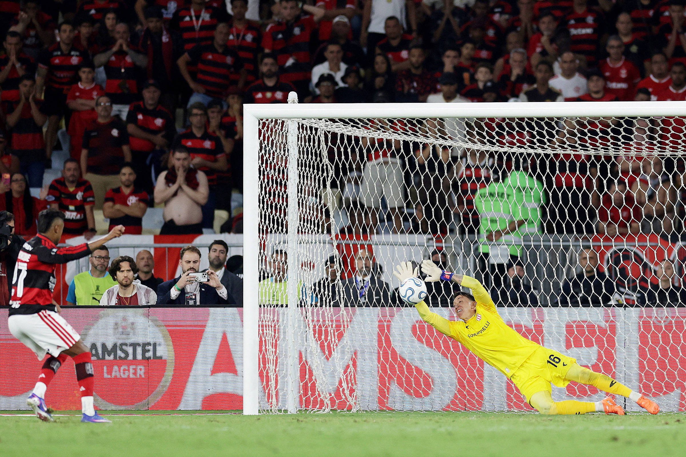 Jogador de futebol do time com camisa listrada vermelha e preta chuta a bola em direção ao gol, enquanto o goleiro vestido de amarelo mergulha para tentar defender. Torcida lota as arquibancadas ao fundo, com muitos vestindo as cores do time atacante.