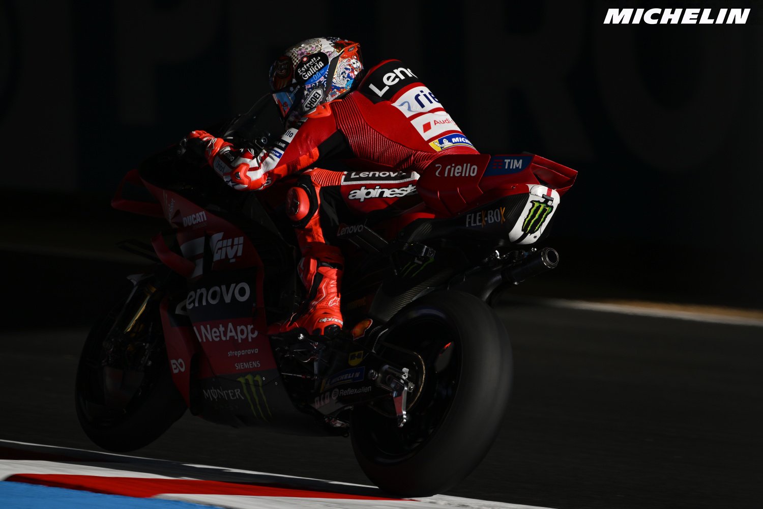 A Ducati MotoGP rider in red and black leathers leans through a corner on his Ducati Desmosedici, with Lenovo and Alpinestars branding visible.
