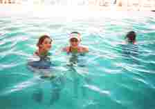 Two women, one in a visor, and a child with snorkeling goggles swim at a pool, smiling at the camera