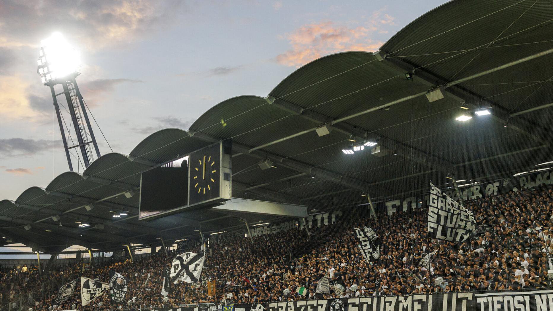 Ein Fußballstadion mit Flutlichtmast und überdachten Rängen, in dem zahlreiche Fans Fahnen schwenken und Banner wie „Grazer Sturmflut“ halten.