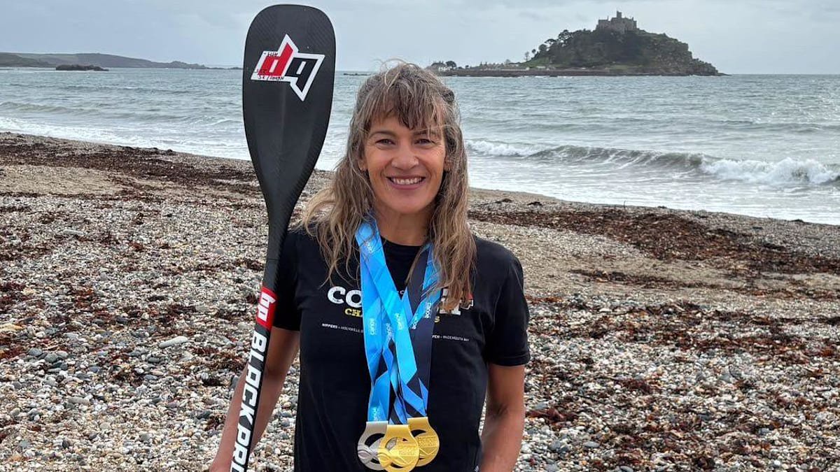 Stand-up paddleboarder Ginnie Betts from Cornwall is stood on a pebble beach with her ore and medals around her neck. She is looking straight at the camera and smiling. The sea and St Michael's Mount is behind her in the background.  