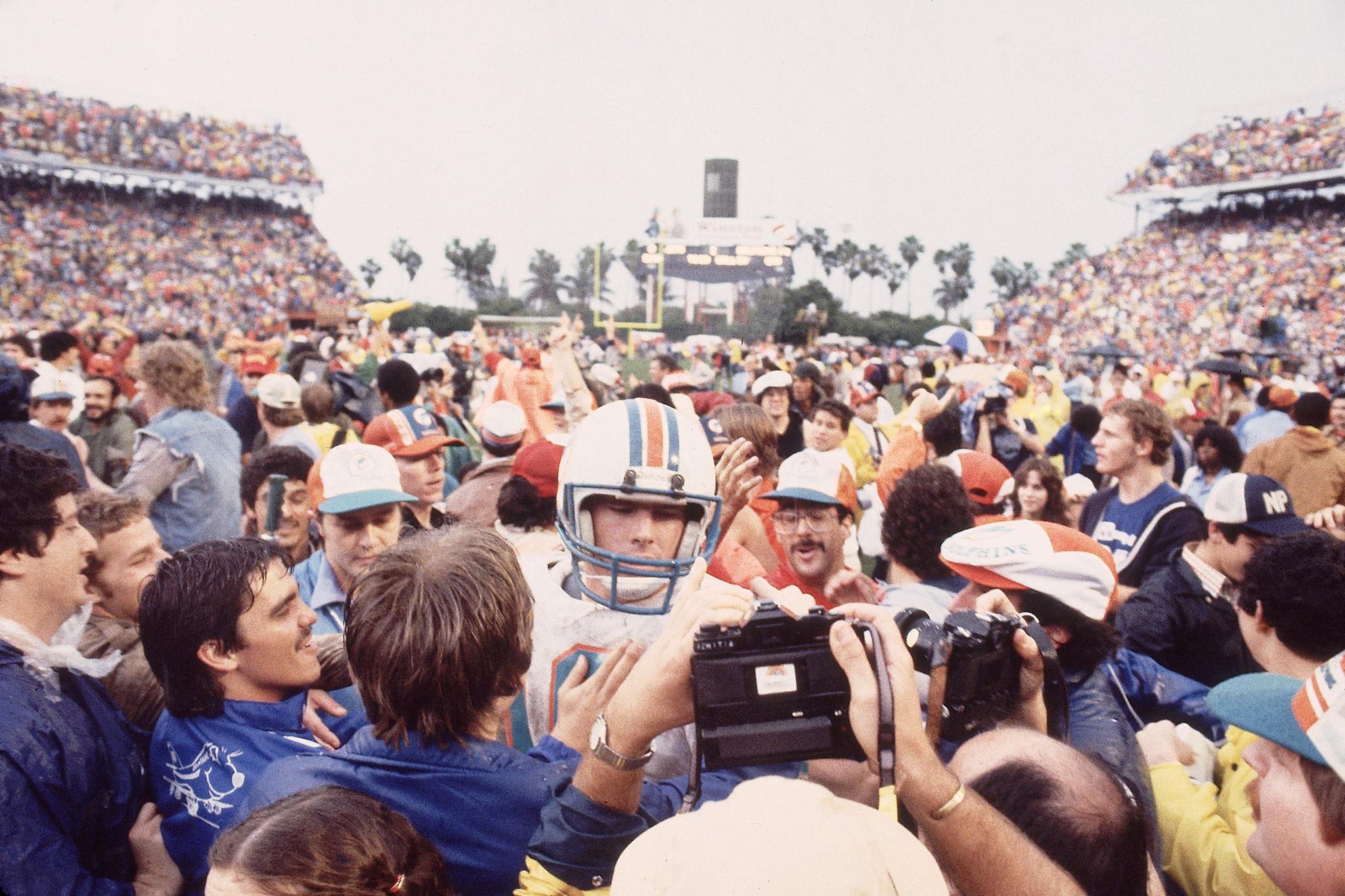 Miami Dolphins quarterback surrounded by cheering fans and photographers on the field.