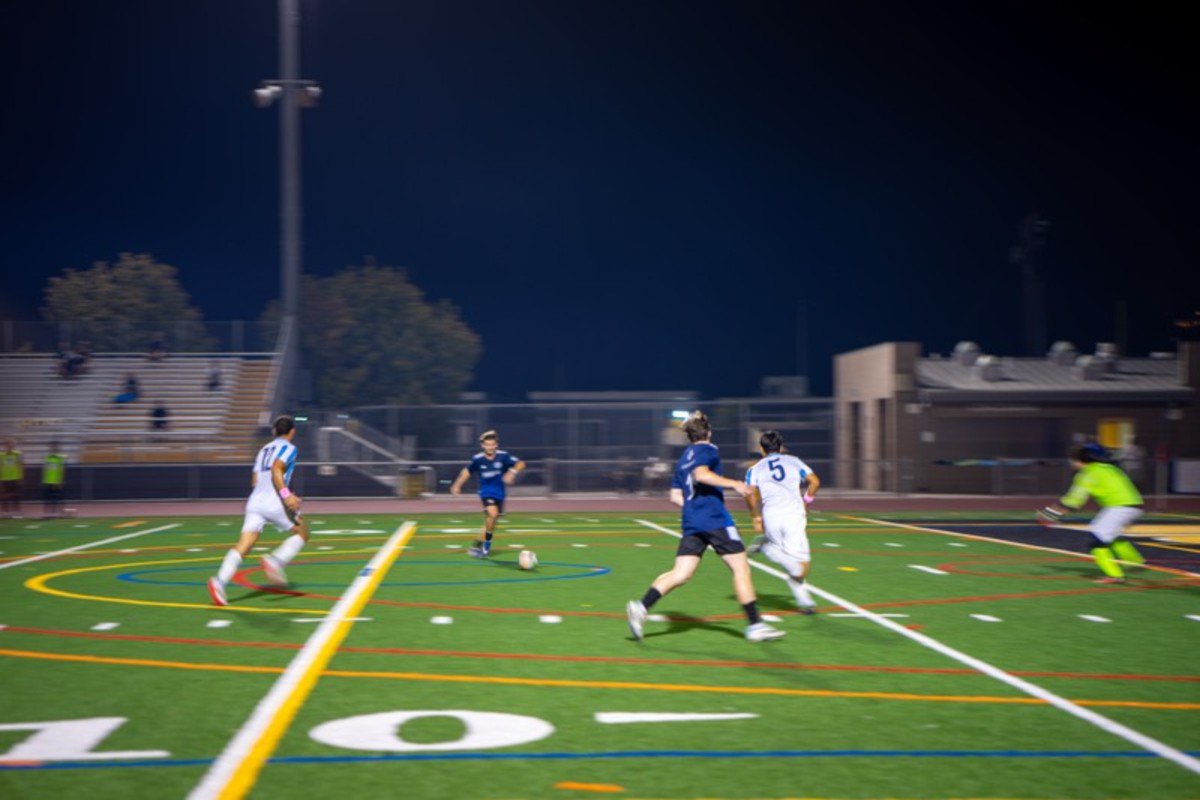 Two opponents on the soccer pitch near a player at their center who has possession of the soccer ball. 