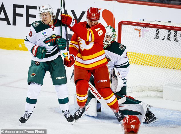 Minnesota Wild's Bobby Brink, left, is hit by the puck as he battles with Calgary Flames' Ben Jones, center, as Wild goalie Jesper Wallstedt, right, reactss during the second period of an NHL hockey game in Calgary, Alberta, Thursday, Oct. 22, 2026. (Jeff McIntosh/The Canadian Press via AP)