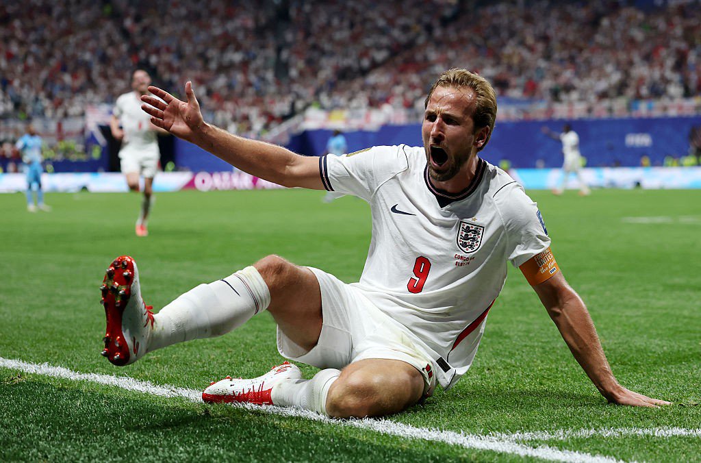 Jogador de futebol Harry Kane, loiro, barba rala, camisa branca número 9 da Inglaterra, calção branco e chuteiras brancas com detalhes vermelhos, sentado no gramado verde com a boca aberta e braços estendidos, celebrando um gol. Um estádio lotado aparece desfocado ao fundo