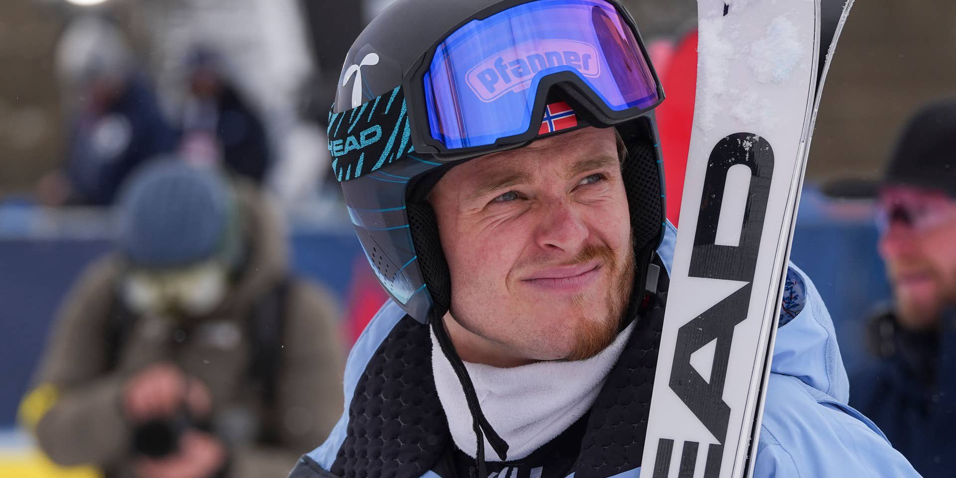 Ein Skifahrer im blauen Anzug hält Ski und trägt Helm mit Skibrille bei einem Wettkampf.