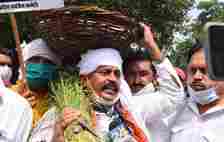 An Indian man carrying a basket on his head and a sheaf of grass in one hand stands in a crowd and speaks into a news microphone.