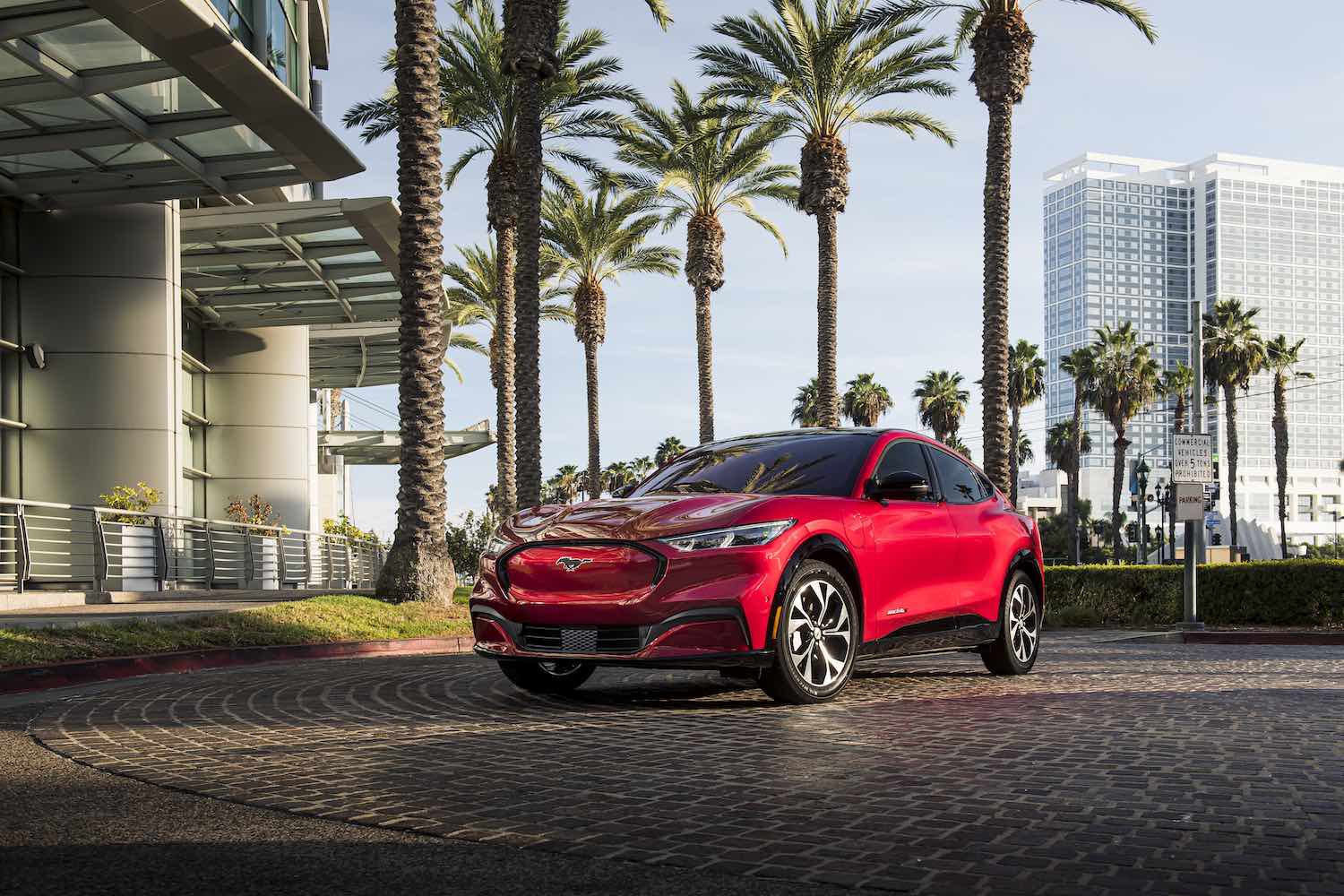 Red Ford Mustang Mach-E crossover parked beneath a row of palm trees in a city.