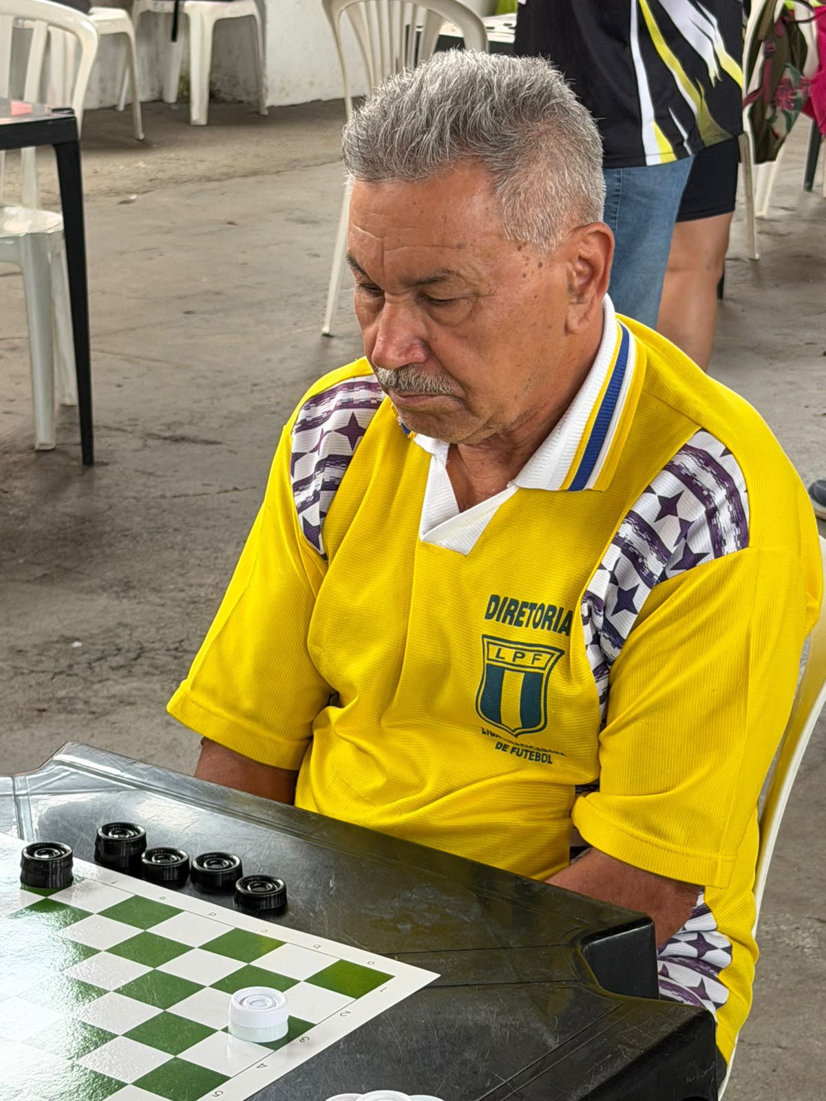 Um homem sénior concentrado a jogar uma partida de damas. Ele está vestido com uma t-shirt amarela da "Diretoria LPF" e encontra-se atento ao tabuleiro com peças pretas e brancas.