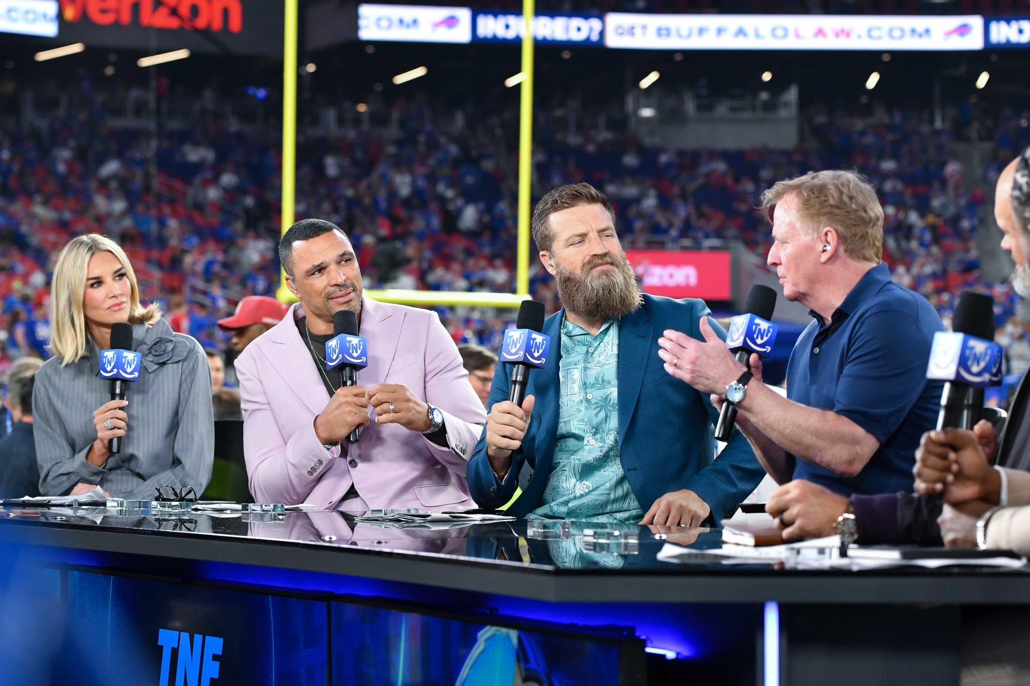 Sports commentators on an NFL broadcast panel with microphones at a desk in a stadium.