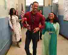 Two actors on a TV set, man holding an award, woman next to him, both smiling, in a school hallway setting