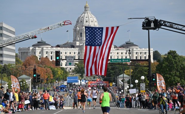 An American flag in the foreground, the St. Paul Capitol in the background, and a crowd of runners and sideline spectators.