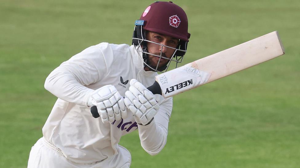 Saif Zaib is wearing cricket whites and a burgundy batters helmet and holding a cricket bat.
