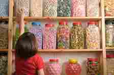 A child in a red shirt reaches for a jar of colorful candies on a shelf filled with various large candy jars