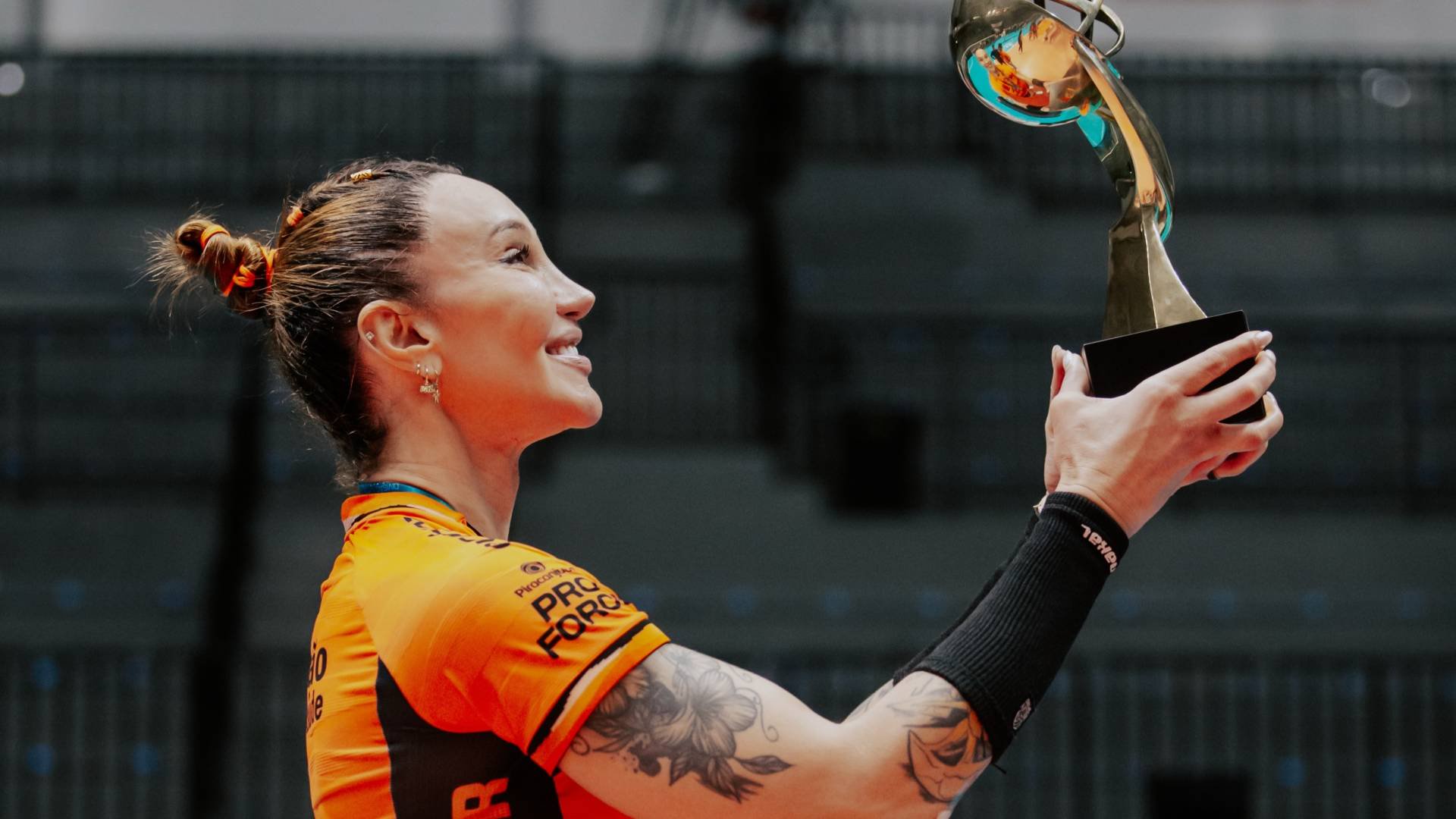 Mulher sorridente de perfil, com cabelo preso e camisa laranja, ergue um troféu dourado com base preta, exibindo tatuagens nos braços, em um ginásio