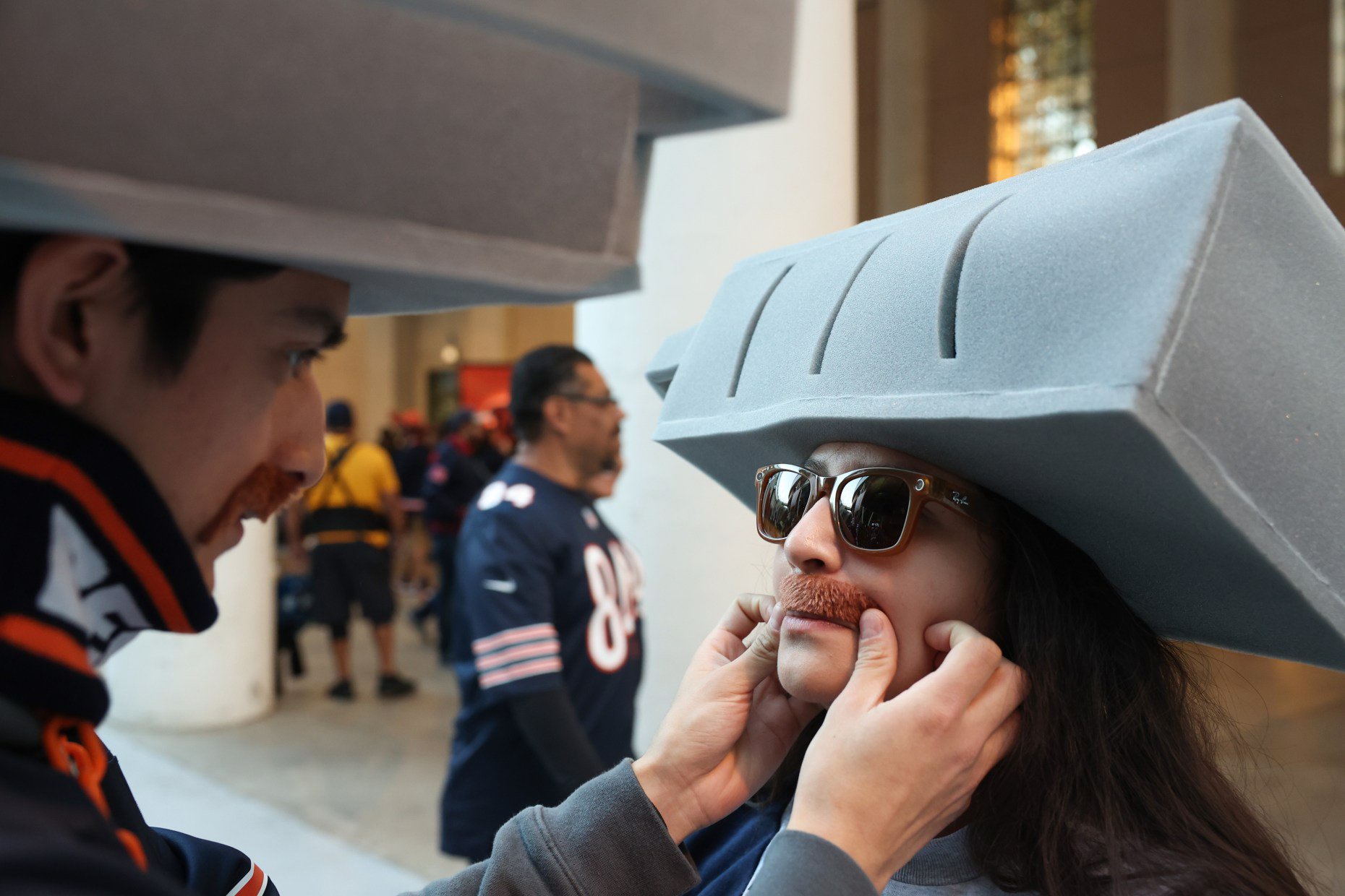 Alessandro Perez places a fake mustache resembling former Chicago Bears head coach and player Mike Ditka on Vanessa Cardenas, given to fans at entry gates, before a Bears-Patriots game at Soldier Field on Oct. 22, 2026, in Chicago. (John J. Kim/Chicago Tribune)