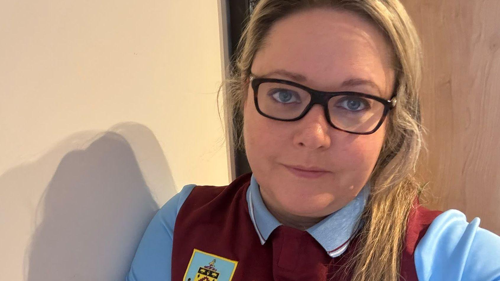 Natalie Bromley has long straight blonde hair and wears dark-framed glasses. She is wearing a claret and blue Burnley FC shirt