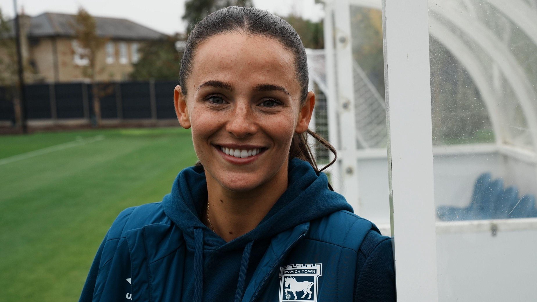 Maria Boswell in a navy Ipswich Town training kit. She is standing in a football dugout and looking directly at the camera.