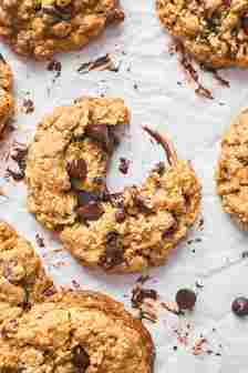 A close up of baked oatmeal chocolate chip cookies with one split apart with chocolate oozing out on a white background