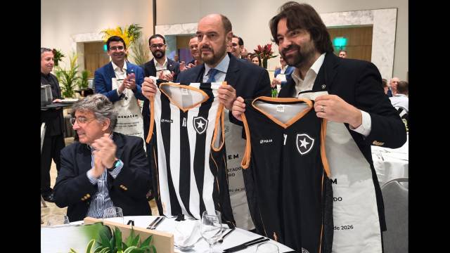 Homens em evento formal, dois sorrindo e segurando camisas de futebol do Botafogo, uma listrada preta e branca e outra preta, com detalhes laranja. Outros homens aplaudem ao fundo, e um homem de óculos e cabelos grisalhos aplaude sentado à mesa, em primeiro plano