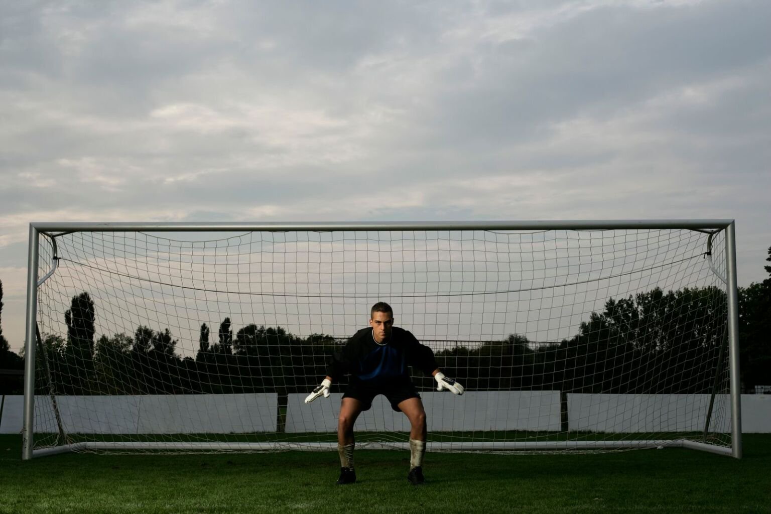 Goalkeeper crouched and ready on his line in front of an empty net at dusk