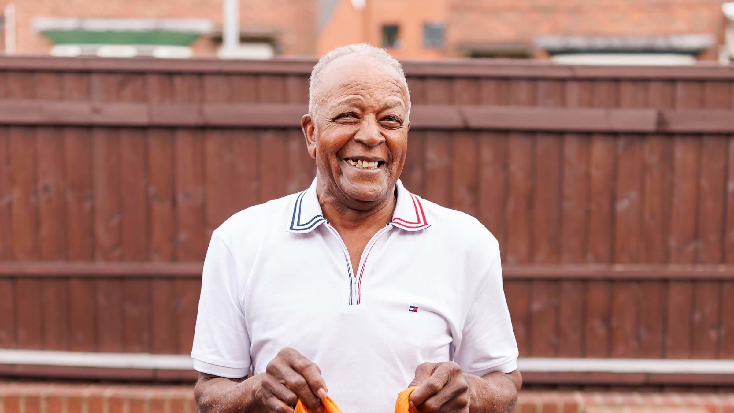 A man in a white polo shirt holding a gold football shirt is smiling widely at the camera. He is standing in front of a brown wooden fence.