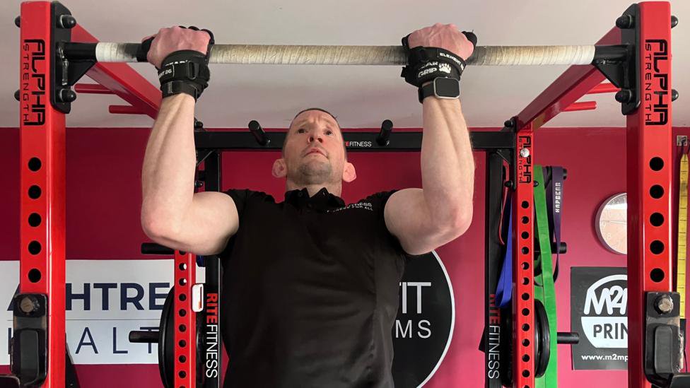 A man wearing a black T-shirt and black wrist bands pulls himself up on a bar in a gym setting.