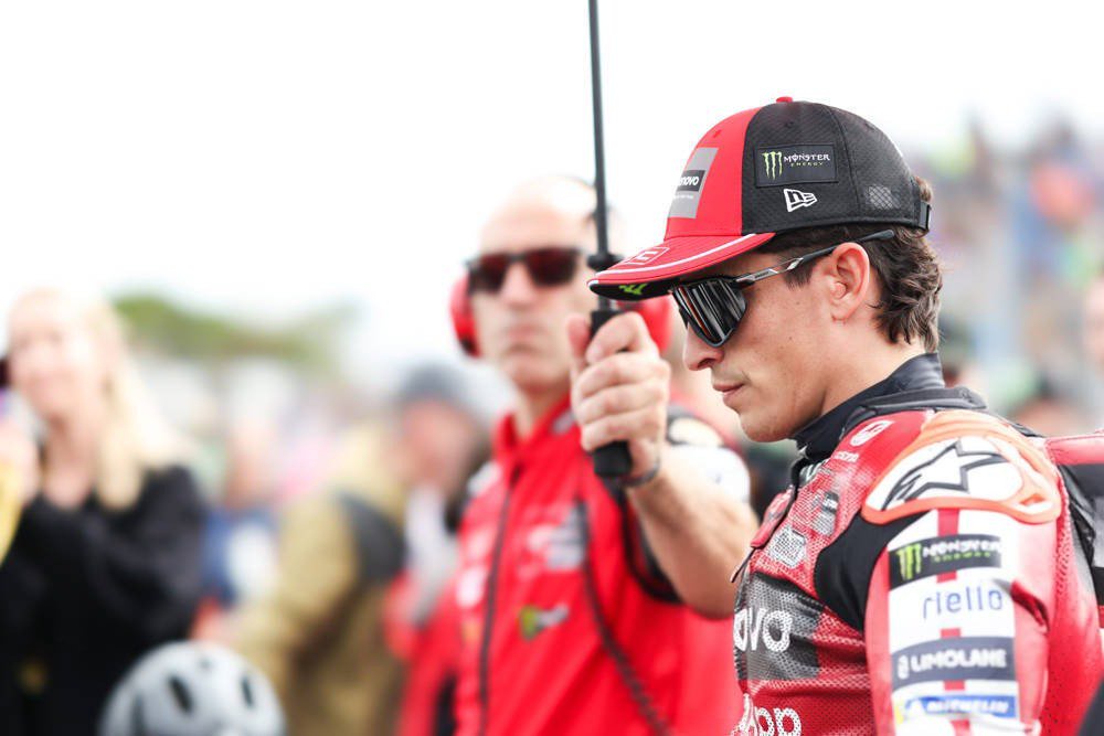 A Ducati rider in red and black leathers and cap holds a radio near a team official in the MotoGP paddock