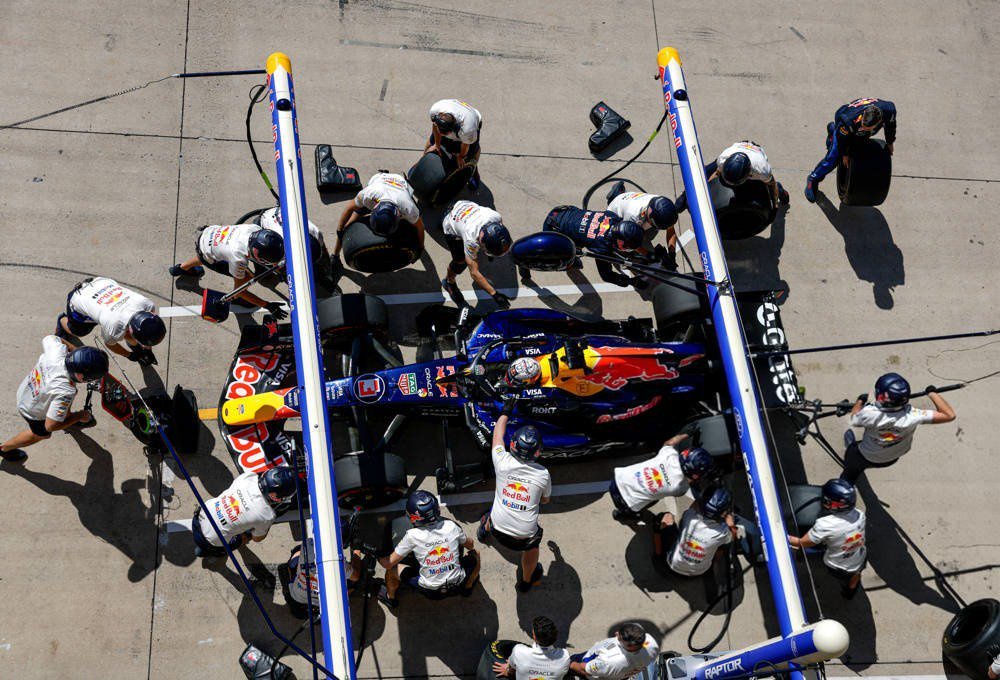 Red Bull Formula 1 pit crew in white and navy kit surrounding a blue Red Bull car during a pit stop, viewed from above.