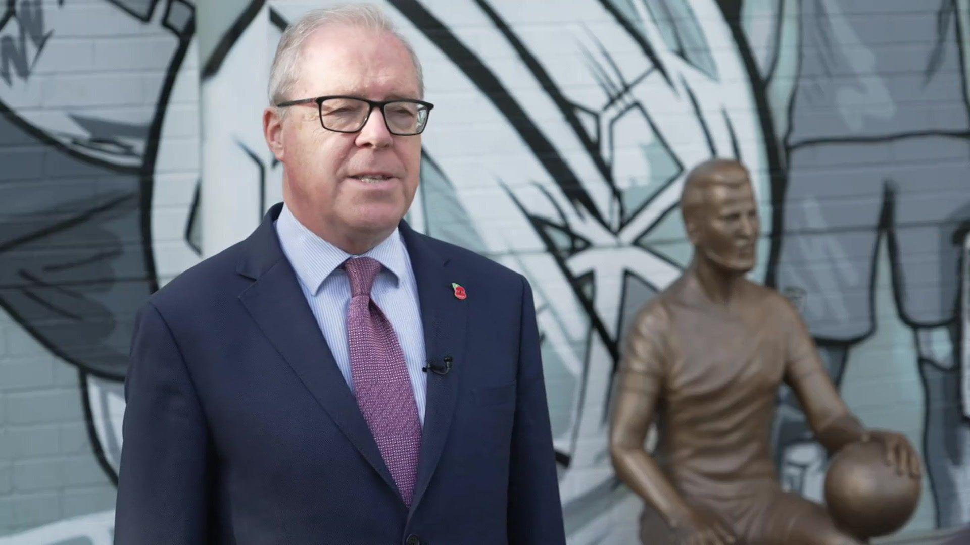 Alex Welsh in a blue suit and purple tie. He has glasses and stands in front of the Harry Kane statue.