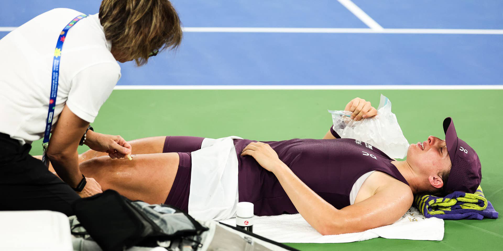 Iga Swiatek liegt auf dem Tennisplatz auf dem Rücken und hält sich einen Eisbeutel auf die Brust, während eine Physio ihren Oberschenkel behandelt.