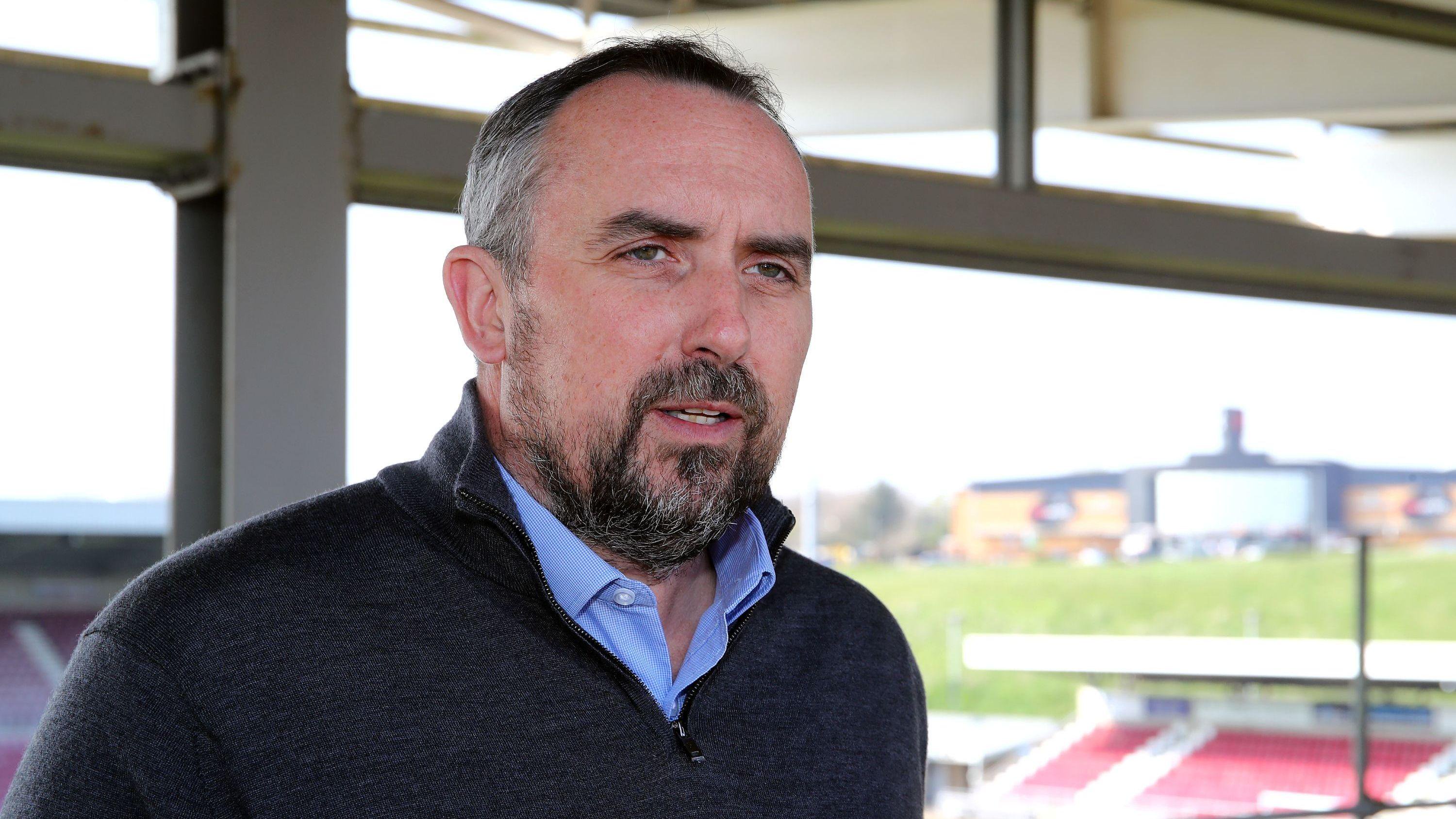 Kelvin Thomas in a blue shirt and dark grey jumper looking slightly off camera. Behind him is a football stadium stand.