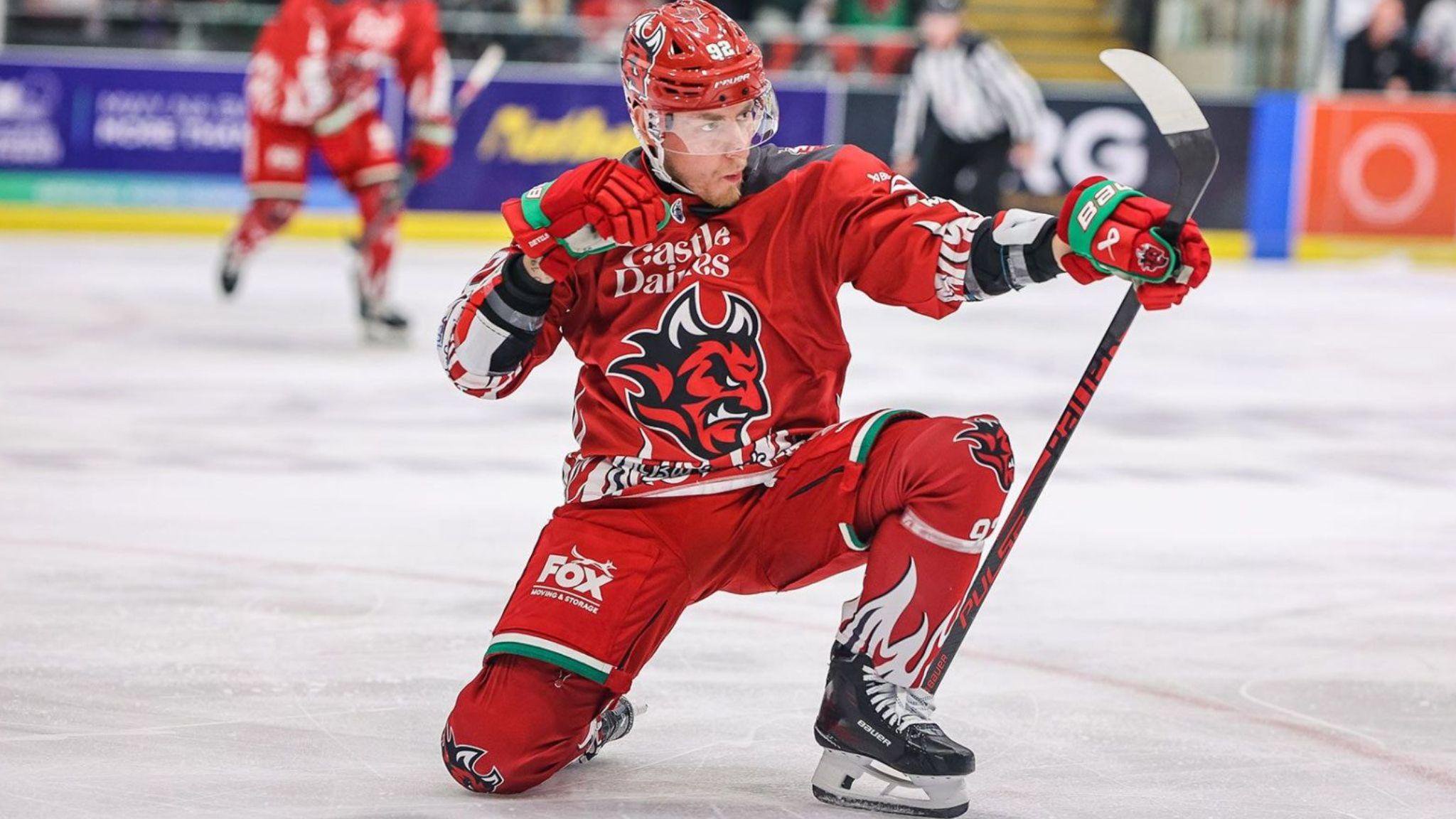 Josh MacDonald, in the red kit of Cardiff Devils, celebrates with one knee on the ice and using his stick to pose as if firing a bow