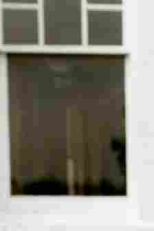 A blurred image of a window with white frames, obscuring any details behind the glass. The window is divided into smaller panes at the top and a larger pane at the bottom. The background appears dark and indistinct. The window is part of a light-colored wall.