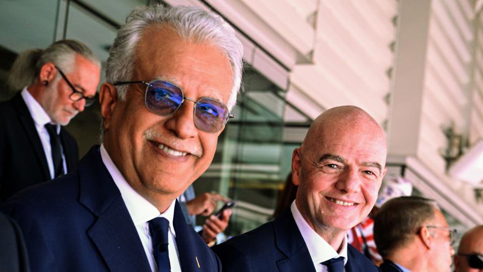 Sheikh Salman bin Ibrahim Al Khalifa and Gianni Infantino stood next to one another in the stands of a football stadium, smiling and looking to their right towards the camera