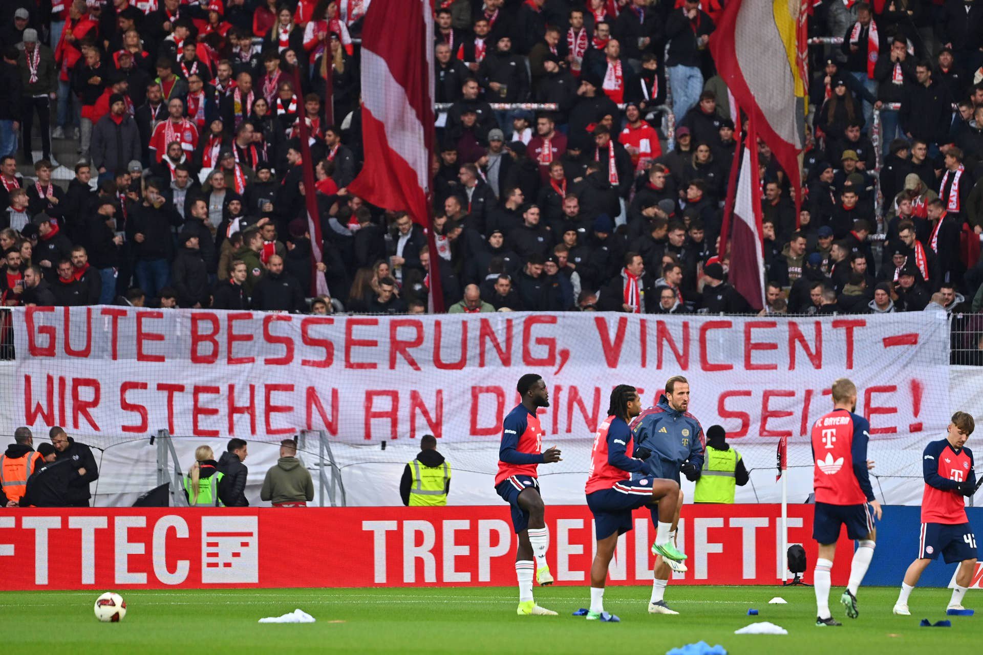 Bayern-Fans halten ein Genesungsbanner für Trainer Vincent Kompany, Spieler wärmen sich auf.