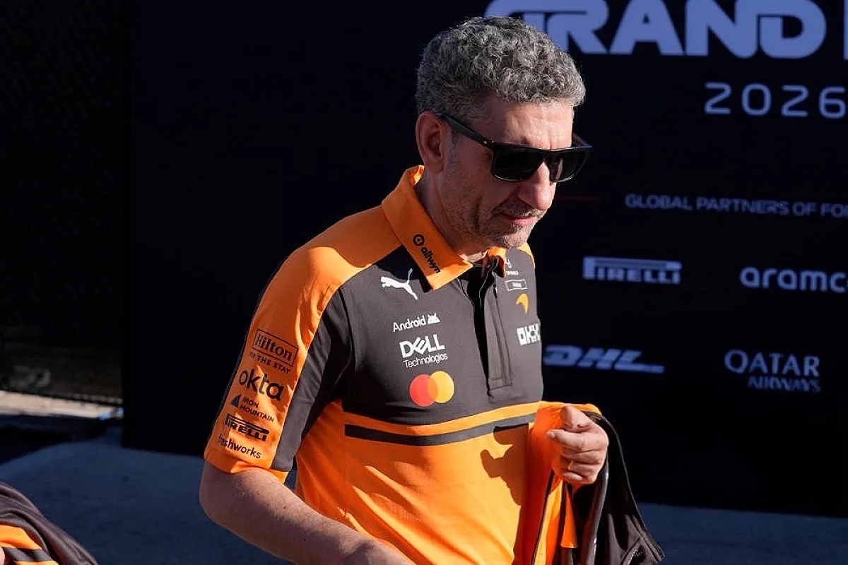 A man in an orange McLaren polo and sunglasses walks past a Grand Prix 2026 backdrop, Formula 1 paddock, sponsors visible