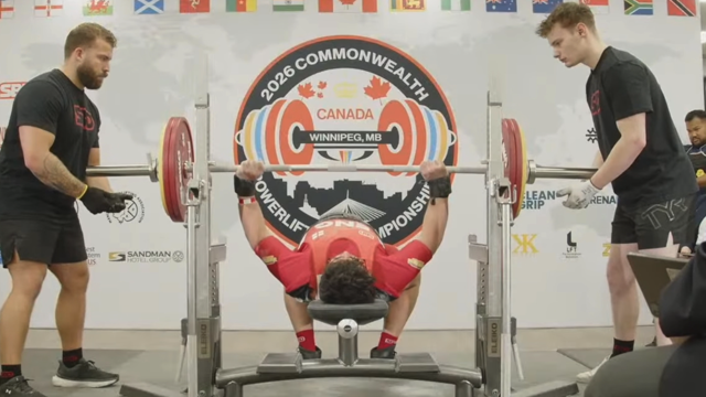 A powerlifter performs a bench press, with two men wearing black T-shirts and shorts standing at either side of the barbell.