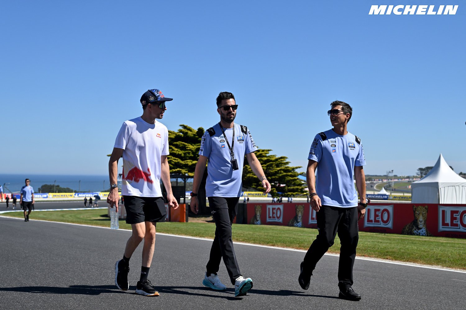Three men in team polo shirts walk along a racetrack past Leo and Michelin branded barriers on a sunny day (MotoGP paddock setting)