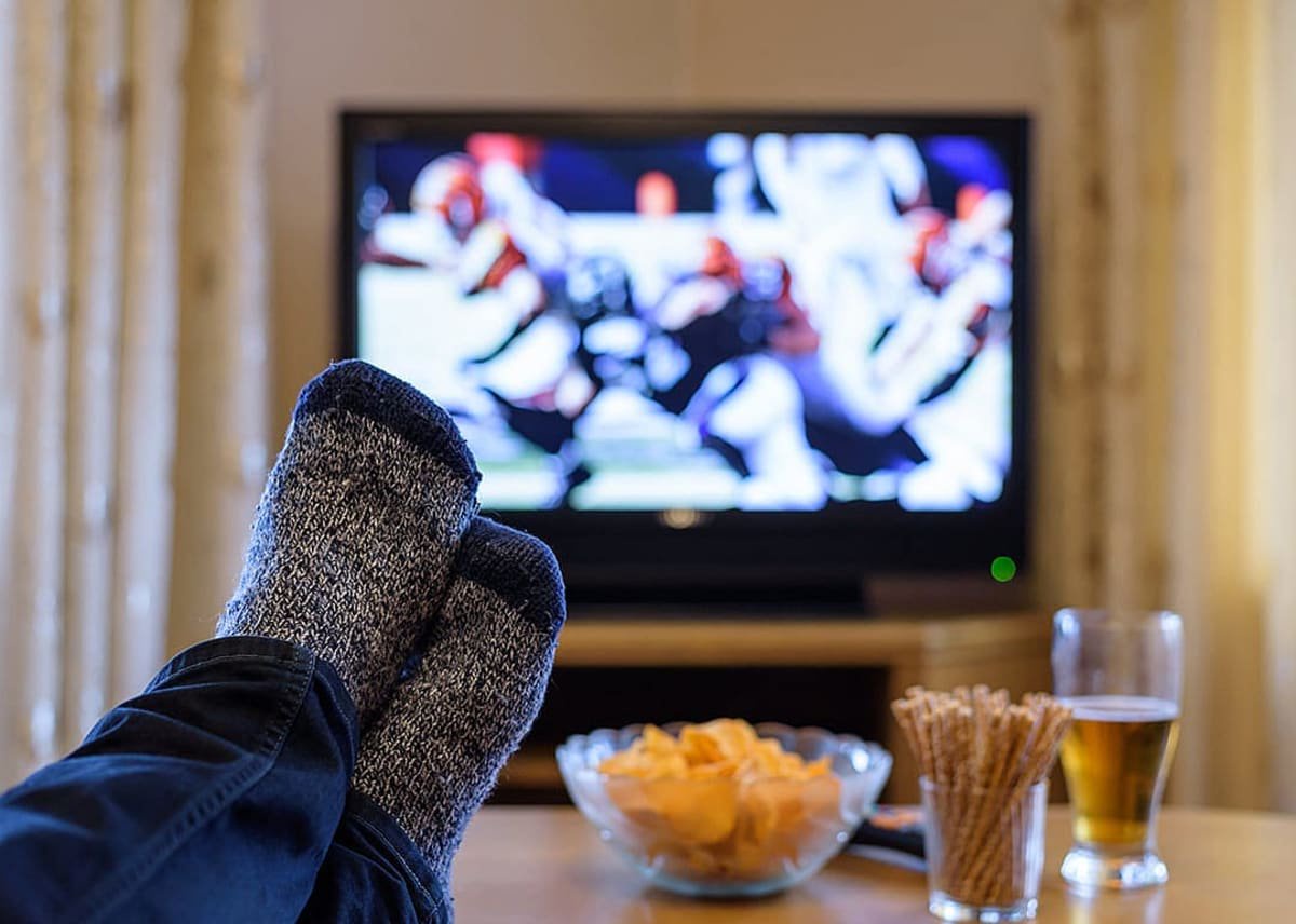A man's feet on a table while watching a football match on a television. - Blackregis // Shutterstock