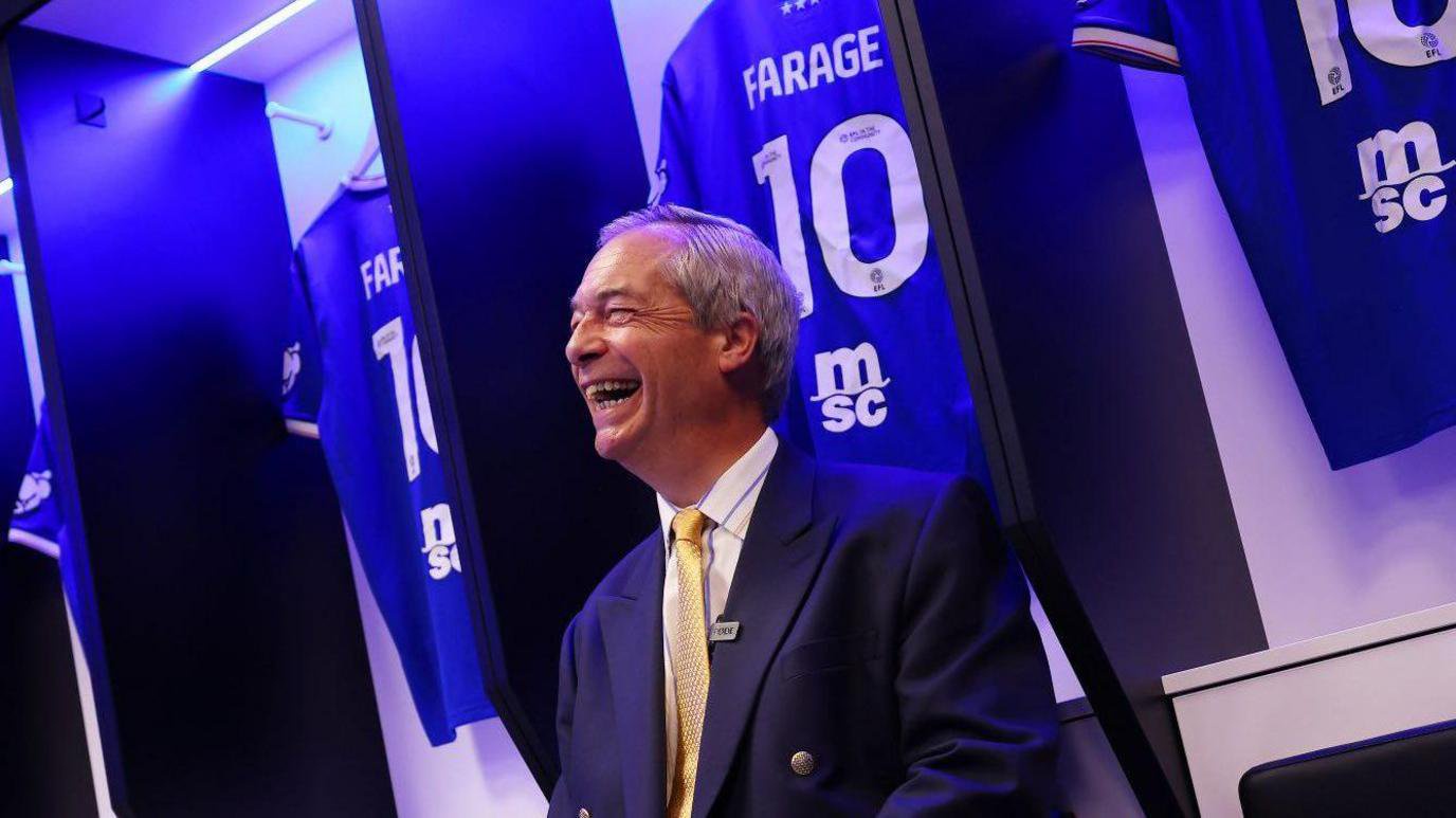 Nigel Farage in a navy suit and a yellow tie sits in Ipswich Town's home dressing room surrounded by personalised 'FARAGE 10' Ipswich shirts. He is smiling and laughing as the picture is taken.