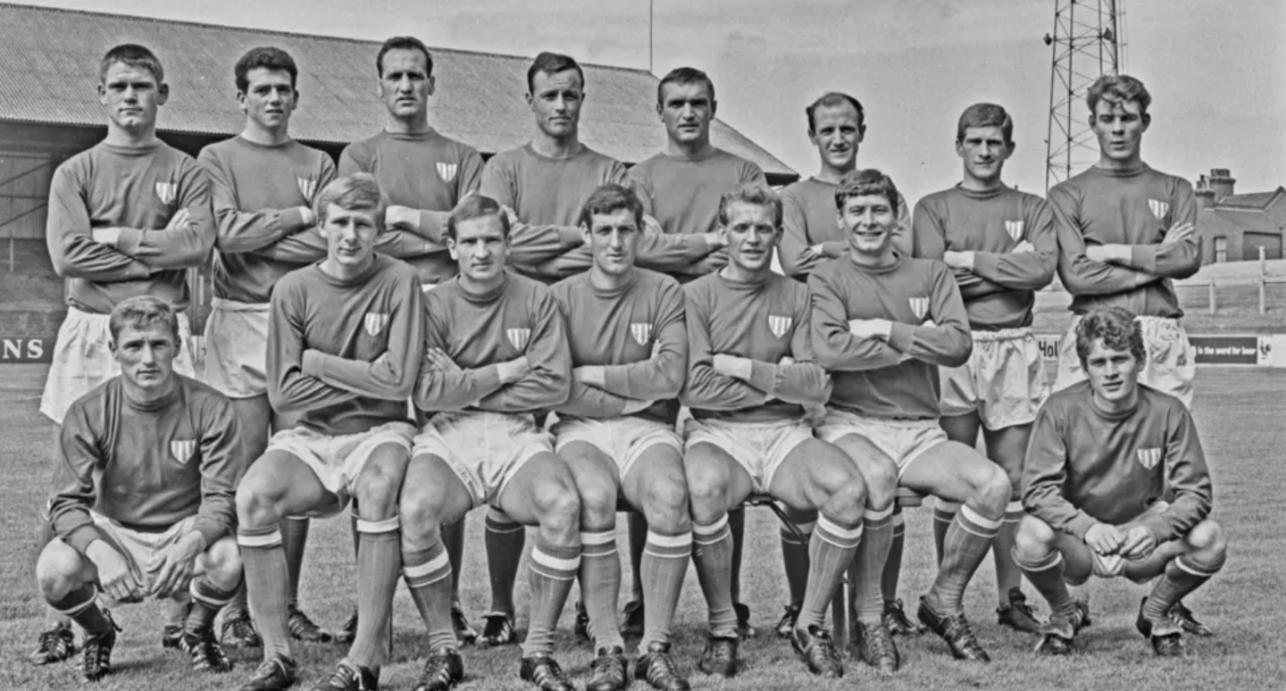 Black and white group photograph of 15 football players. Half are standing up, the rest sitting down. They have their arms folded. They are on the pitch at a football ground. 