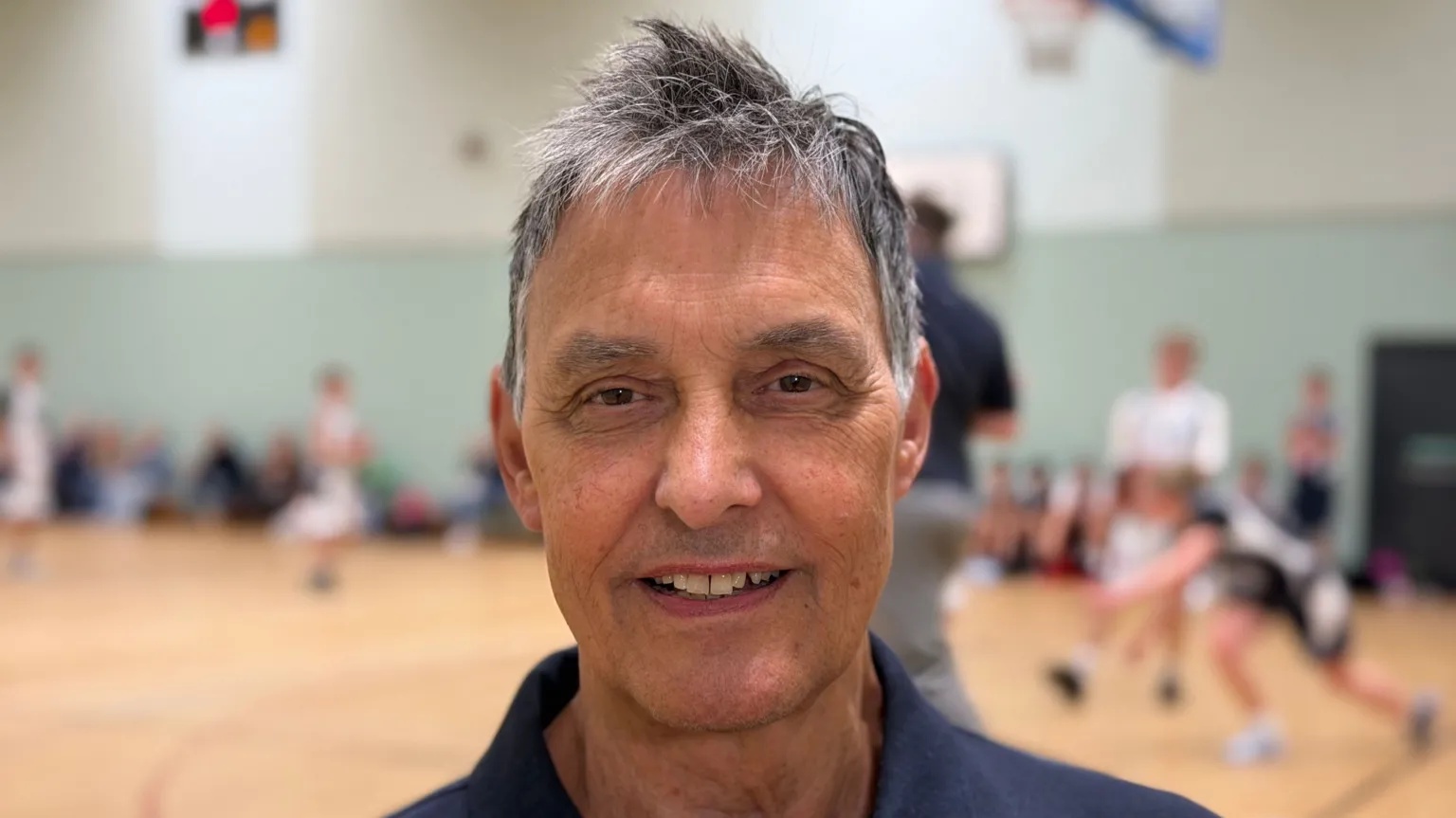 Photo of Roger with a blue top on, standing inside a sports hall. 