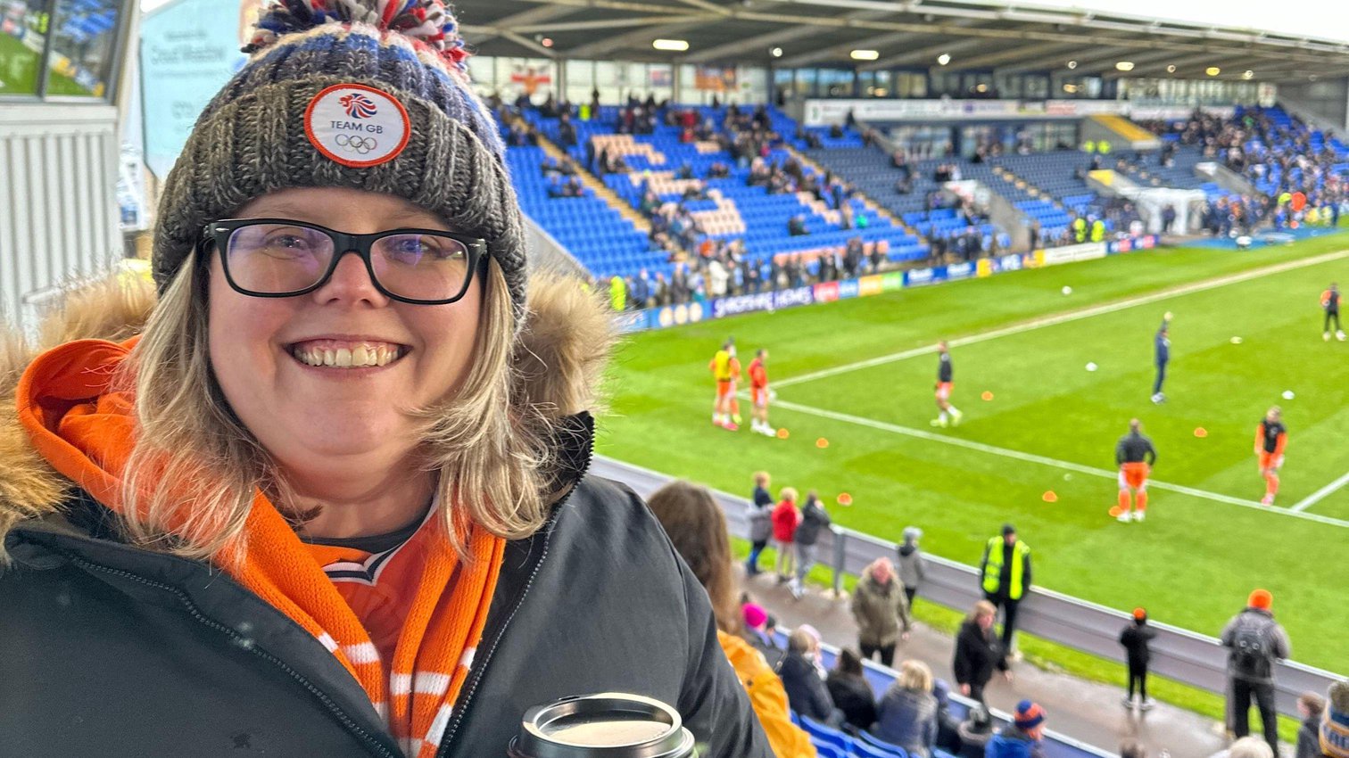 Fiona Martin is wearing a Team GB woolly hat and glasses. She is wearing a bright orange hoodie underneath a dark jacket. In the background is a football stadium.
