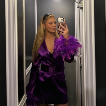 A woman with long blonde hair and sunglasses on her head takes a mirror selfie while wearing a purple satin dress, a belt, and purple feather cuffs on her wrists.