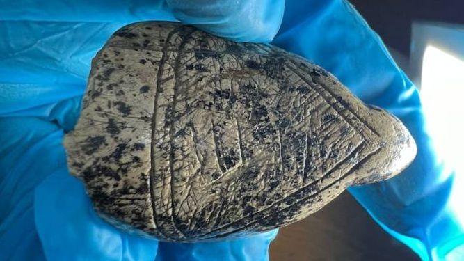A gloved hand holds a small carved piece of mammoth ivory with engraved geometric lines and a weathered, speckled surface.