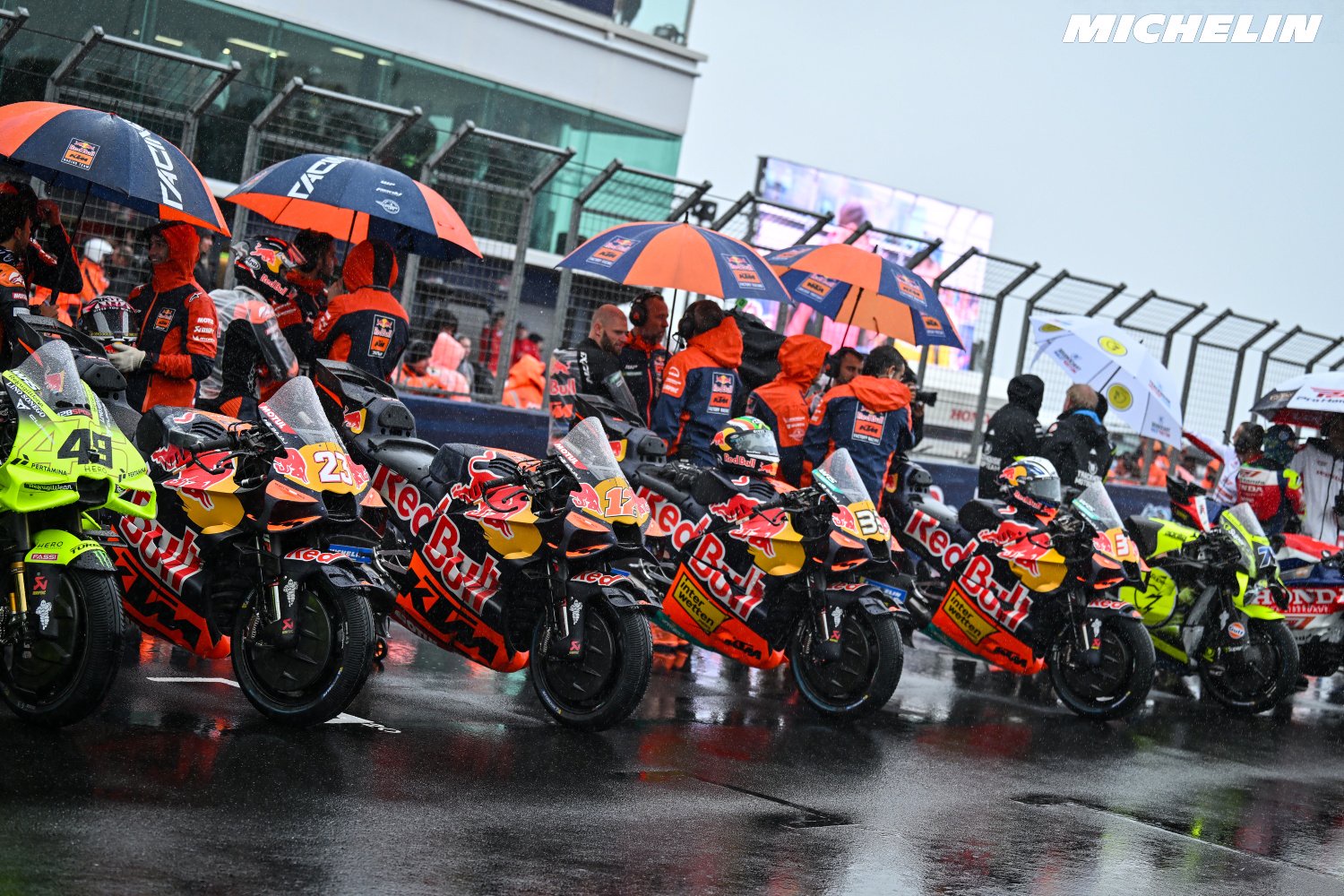 MotoGP riders with Red Bull KTM bikes lined up on a wet grid under umbrellas, while crew in rain gear look on